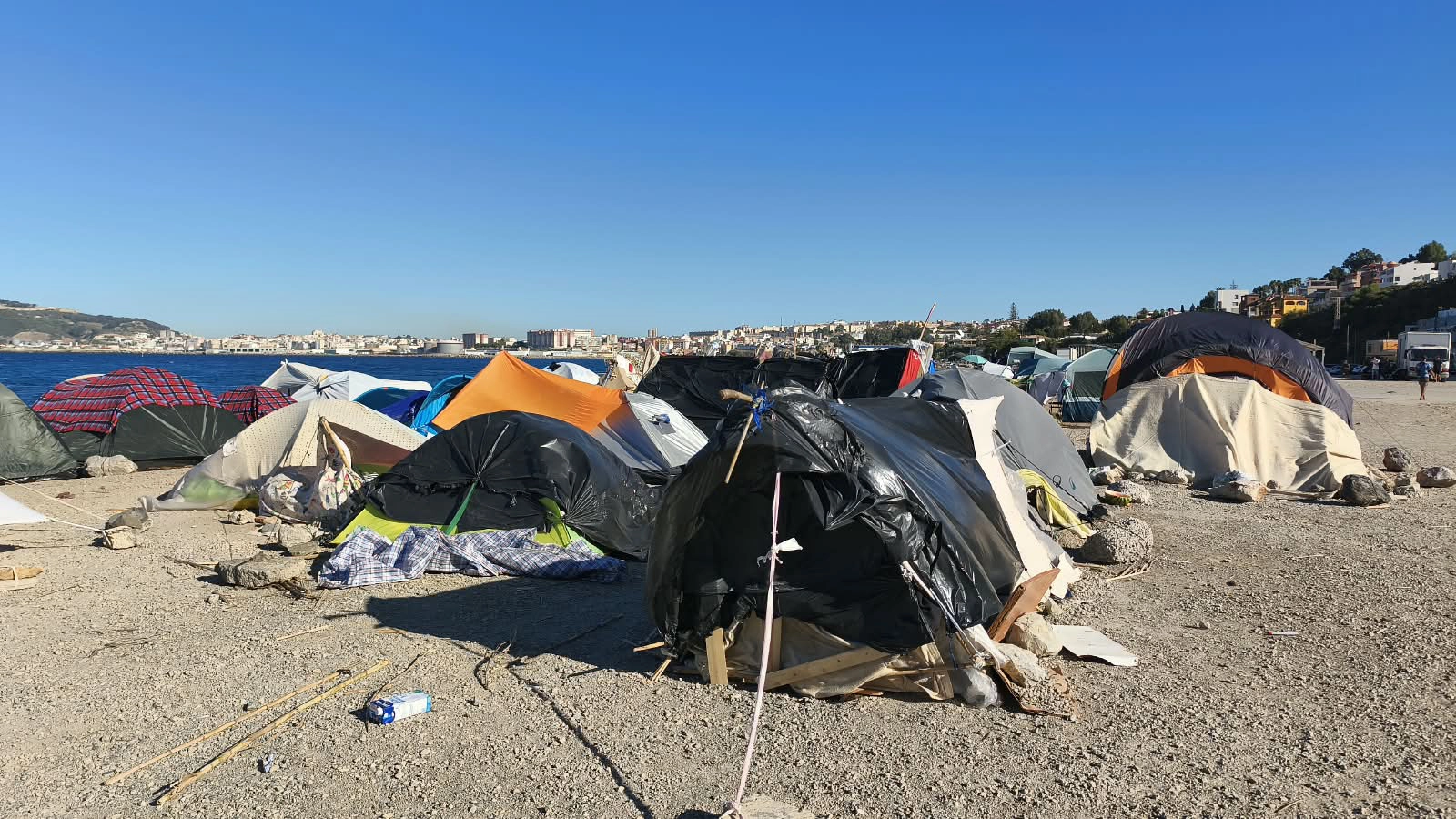 Campamento improvisado de tiendas de campaña y plásticos en la playa de Ceuta, con la ciudad y el mar al fondo.
